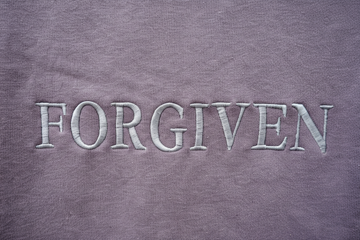 Close-up detail of white tonal embroidered FORGIVEN lettering on dusty mauve crewneck